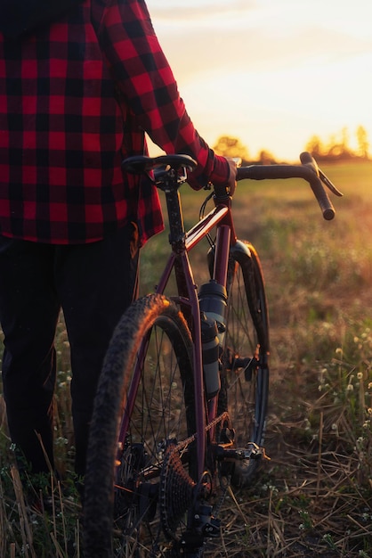 Foto captura aproximada de um ciclista ativo vestindo uma camisa quadriculada segurando uma bicicleta de cascalho em um campo ao pôr do sol