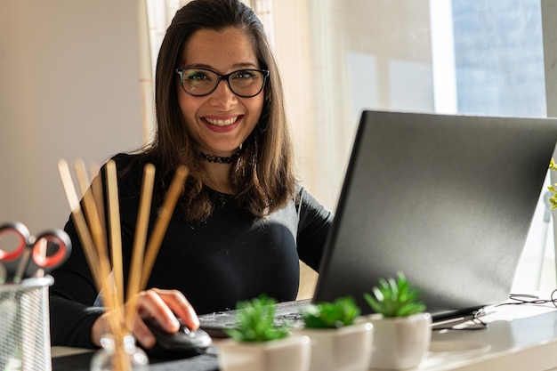 Captura aproximada de uma atraente mulher latina sorridente em óculos trabalhando em um laptop em seu escritório