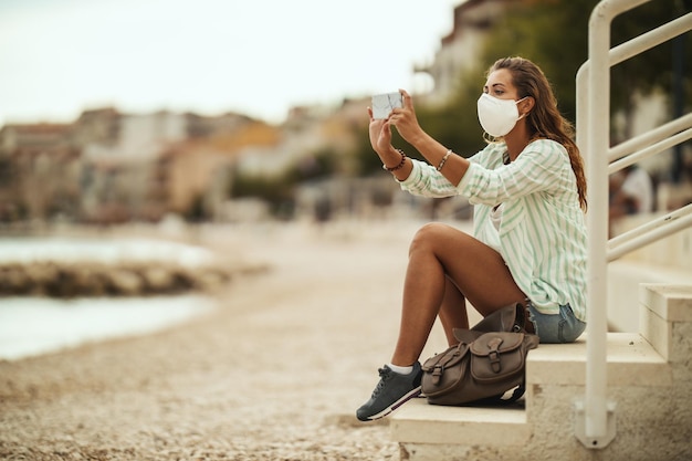 Captura de una atractiva joven feliz que usa una máscara protectora N95 y toma una foto con un teléfono inteligente suyo mientras disfruta de unas vacaciones en la playa durante el COVID-19.