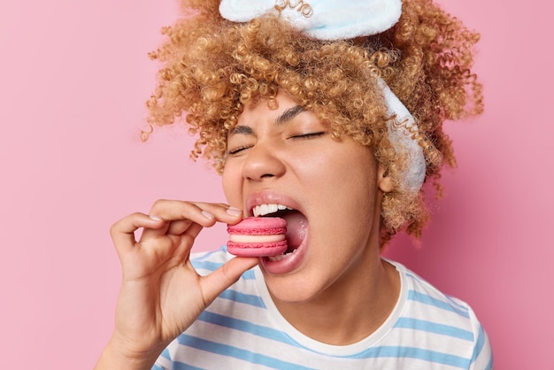 Captura de cabeza de una joven de cabello rizado que come dulces y deliciosos macarrones que prefiere alimentos poco saludables que contienen azúcar y usa una camiseta a rayas con banda para la cabeza aislada sobre un fondo rosa Concepto de confitería