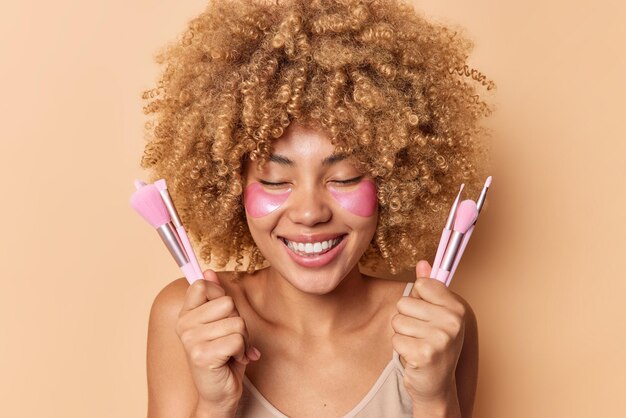 Captura de cabeza de una mujer europea de pelo rizado positivo que se ríe felizmente sostiene cepillos cosméticos para aplicar la base que va a maquillarse después de que los tratamientos para el cuidado de la piel usen parches de hidrogel debajo de los ojos