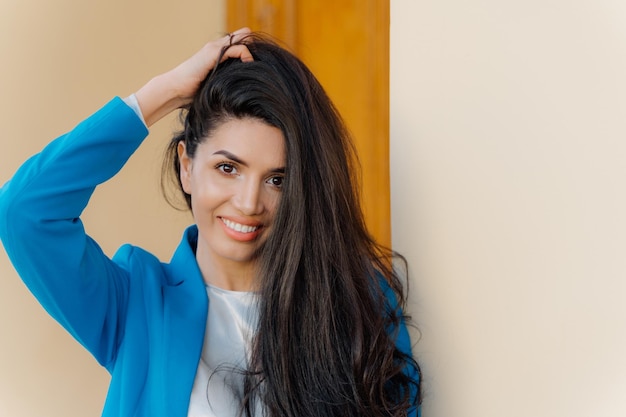 Captura de cabeza de mujer morena feliz con maquillaje de sonrisa suave viste ropa elegante contenta después de una exitosa reunión de negocios mira a la cámara mantiene la mano en el cabello La señora alegre posa con ropa formal