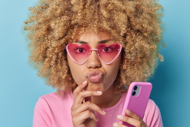 Captura de cabeza de una mujer seria y atenta con cabello rizado que mantiene los labios doblados, mira directamente a la cámara, usa el teléfono inteligente, piensa en descargar una nueva aplicación, usa gafas de sol y una camiseta informal aislada en azul