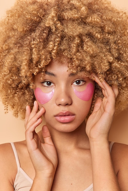 Captura de cabeza de una mujer seria de cabello rizado que toca la cara suavemente, se ve segura de sí misma, aplica parches de hidrogel rosa debajo de los ojos para el rejuvenecimiento de la piel, se somete a tratamientos de belleza y quiere lucir joven