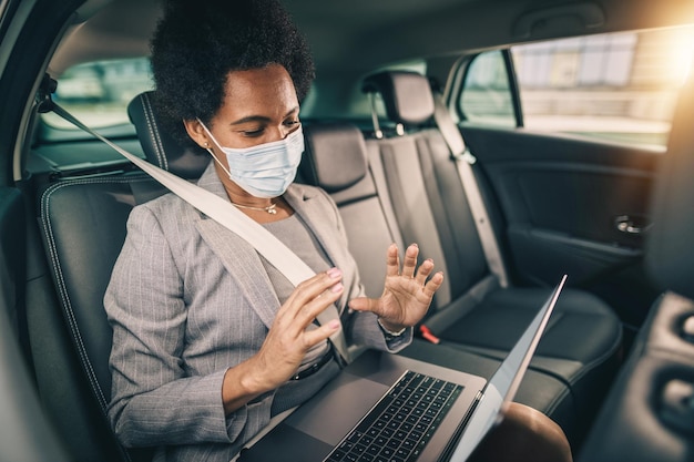 Captura de una exitosa mujer negra con máscara protectora que hace una videollamada en una laptop mientras está sentada en el asiento trasero de un camión en su viaje de negocios durante la pandemia de COVID-19.