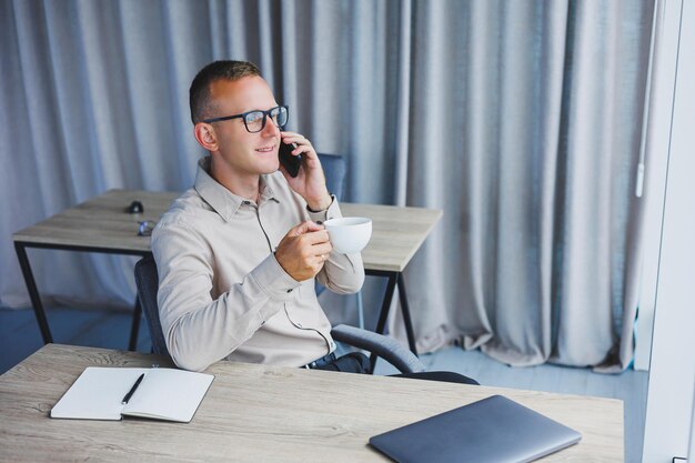 Captura interior de un alegre trabajo independiente masculino barbudo en la gestión distante de las finanzas a través de un teléfono inteligente sentado en una mesa de madera que trabaja con documentación tiene una expresión feliz Tecnología de trabajo de personas