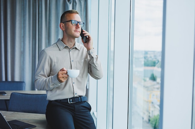 Captura interior de un alegre trabajo independiente masculino barbudo en la gestión distante de las finanzas a través de un teléfono inteligente sentado en una mesa de madera que trabaja con documentación tiene una expresión feliz Tecnología de trabajo de personas