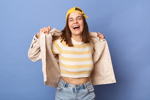 Captura interior de una hermosa adolescente alegre con gorra de béisbol y chaqueta posando aislada sobre fondo azul mantiene la mano en su ropa regocijándose riendo divirtiéndose