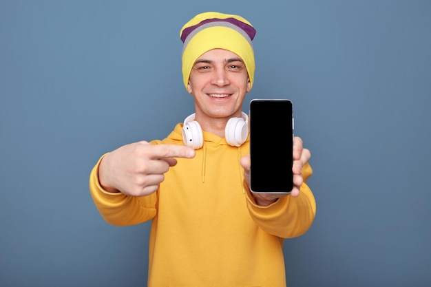 Captura interior de un hombre alegre y encantado con sombrero de beanie y capucha amarilla casual parado aislado sobre fondo azul apuntando a un teléfono inteligente con espacio de copia de pantalla vacío para publicidad