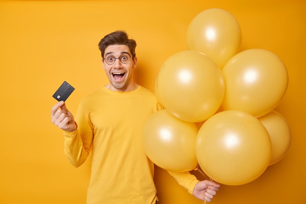 Captura interior de un hombre guapo que exclama en voz alta sostiene una tarjeta de crédito y globos inflados usa un saltador casual celebra cumpleaños aislado sobre fondo amarillo hace el pago en línea Concepto de vacaciones