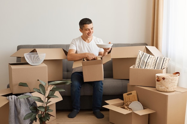 Captura interior de un hombre optimista positivo con camiseta blanca y jeans sentado en un sofá rodeado de cajas de cartón, desempacando paquetes, mudándose a una nueva casa.