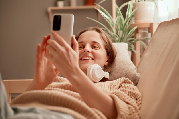 Captura interior de una joven adulta atractiva que usa audífonos usando un teléfono inteligente mientras está acostada en el sofá sonriendo felizmente disfrutando del tiempo libre en el fin de semana navegando por Internet