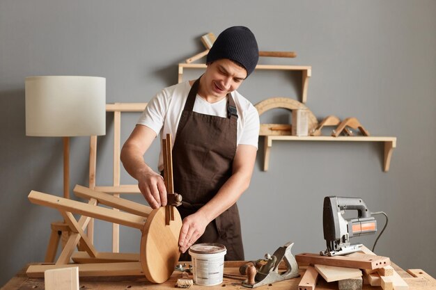 Captura interior de un joven adulto caucásico carpintero con camiseta blanca, gorra negra y delantal marrón trabajando haciendo una silla de madera disfrutando de su hobby y trabajo