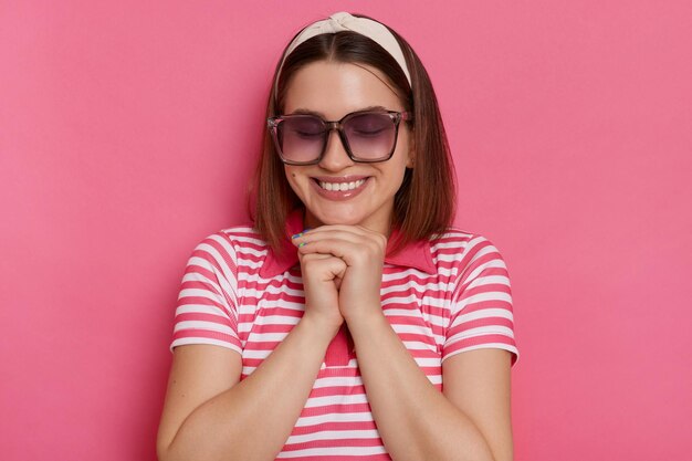 Captura interior de una linda y sonriente mujer encantadora que usa camiseta a rayas y gafas de sol mantiene los ojos cerrados manteniendo las manos debajo de la barbilla posando aisladas sobre un fondo rosado