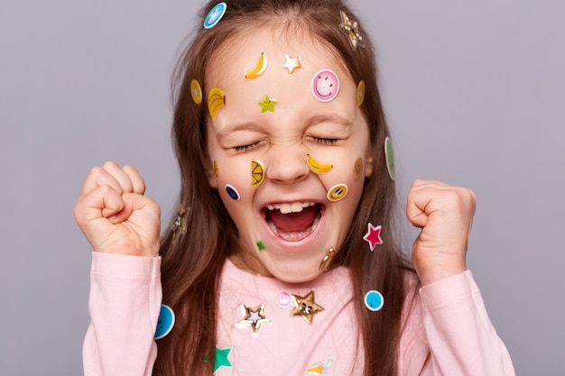 Captura interior de una linda y sonriente niña de cabello castaño cubierta de pegatinas posando aislada sobre un fondo gris con los puños apretados celebrando su éxito