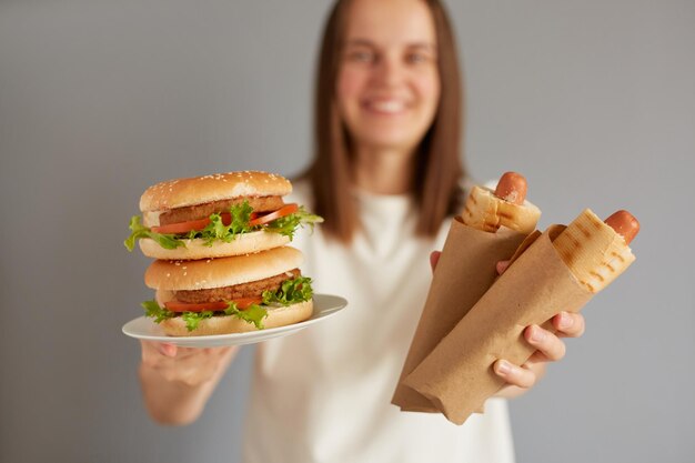 Captura interior de una mujer amigable positiva sosteniendo un perro caliente y una hamburguesa usando una camiseta blanca posando aislada sobre un fondo gris que ofrece comida chatarra sabrosa para ti