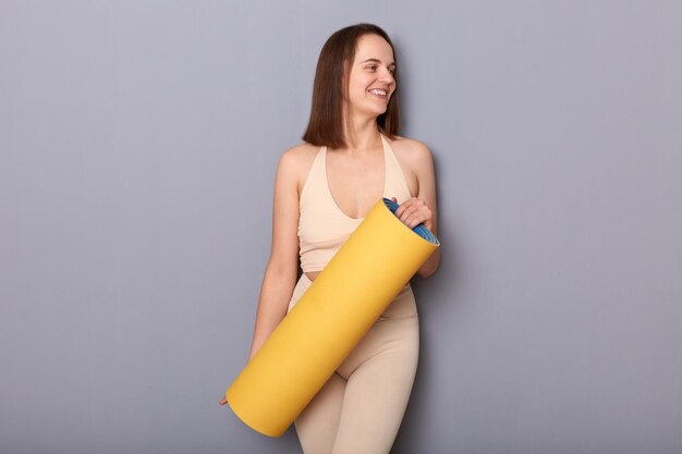 Captura interior de una mujer atlética alegre y sonriente que mira con una colchoneta de fitness enrollada en buena forma física tiene entrenamiento deportivo todos los días espacio libre