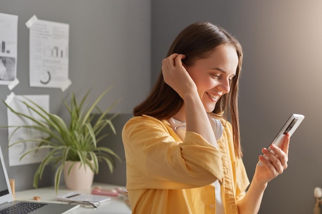 Captura interior de una mujer caucásica sonriente y feliz con cabello castaño y camisa amarilla posando en la oficina y usando un teléfono celular mientras lee un mensaje o publica un mensaje en las redes sociales