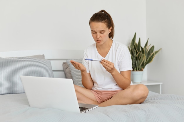 Captura interior de una mujer disgustada con pantalones blanco y pantalones cortos sentada en la cama frente a una computadora portátil sosteniendo una prueba de embarazo con resultado negativo, expresando tristeza, se ve confundida.