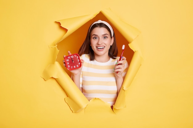 Captura interior de una mujer emocionada, feliz y alegre, con camisa a rayas y banda para el cabello sosteniendo un despertador y un cepillo de dientes, despierta y está lista para hacer procedimientos que rompen el fondo de papel amarillo