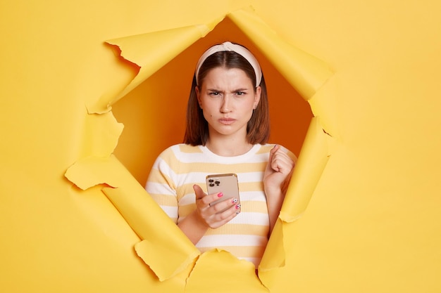 Captura interior de una mujer enfadada y disgustada con un pantalones a rayas mirando a través de un avance de fondo amarillo sosteniendo un teléfono móvil y mirando la cámara con ira y odio