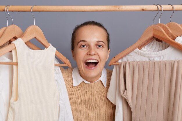 Captura interior de una mujer extremadamente feliz, emocionada y sorprendida, parada cerca de la ropa colgada en el estante, vendiendo muchos atuendos de moda, regocijándose de su éxito, gritando de felicidad mirando a la cámara