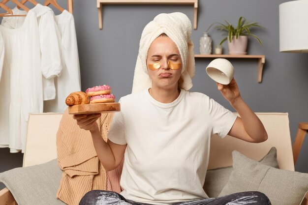 Foto captura interior de una mujer molesta que usa pantalones grises de camiseta blanca y una toalla en la cabeza necesita energía sosteniendo dulces y una taza vacía sentada tosiendo con ropa colgada en el estante en el fondo