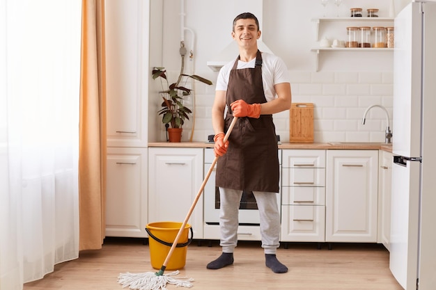 Captura interior de una mujer positiva sonriente que usa delantal marrón y guantes de goma sosteniendo un piso de lavado de mopa en la cocina mirando a la cámara de buen humor mientras hace las tareas del hogar