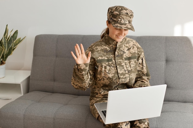 Captura interior de una mujer soldado sonriente y satisfecha con uniforme de camuflaje y gorra sentada en un sofá y trabajando en una laptop con una videollamada saludando con la mano