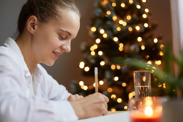 Captura interior de una mujer sonriente y atractiva sentada en el escritorio y escribiendo en un organizador de cuadernos de papel con un árbol de Navidad en el fondo haciendo una lista de deseos para las vacaciones de Año Nuevo