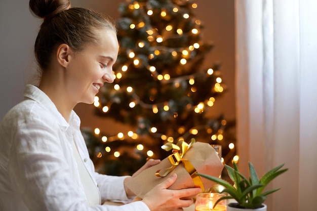 Captura interior de una mujer sonriente con camisa blanca sentada en la mesa y sosteniendo un regalo de navidad con un lazo dorado en el fondo del árbol de navidad con luces que expresan felicidad y emociones positivas