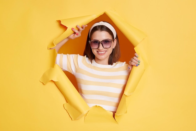 Captura interior de una mujer sonriente y encantada que usa una banda para el cabello de camiseta a rayas y gafas de sol que se abren paso a través de un agujero de papel en una pared amarilla mirando a la cámara con expresión positiva