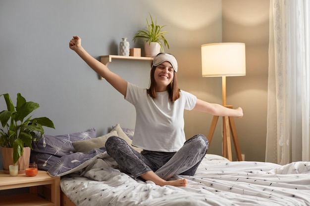 Captura interior de una mujer sonriente y satisfecha que usa una máscara para dormir sentada en la cama en un dormitorio acogedor y estirando los brazos despertándose de buen humor disfrutando la mañana del fin de semana