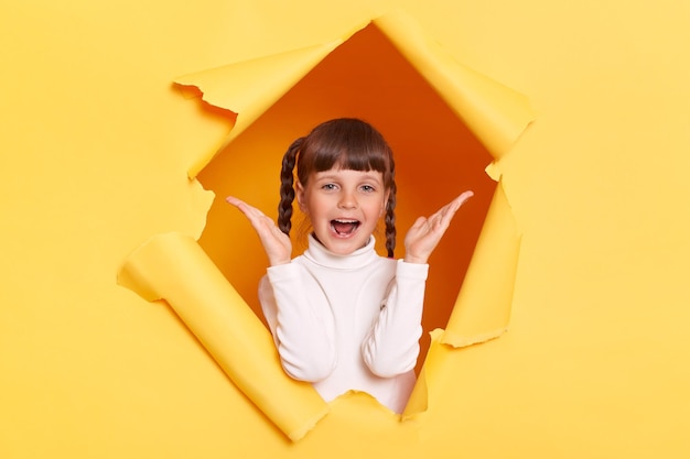 Captura interior de una niña caucásica asombrada y positiva con trenzas con cuello de tortuga blanco posando en un agujero rasgado de una pared de papel amarillo que ve algo sorprendente al ser sorprendida con los brazos levantados