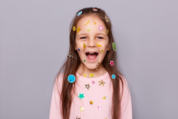 Captura interior de una niñita de cabello castaño extremadamente feliz y emocionada cubierta con pegatinas gritando con expresión alegre regocijándose posando aislada sobre un fondo gris