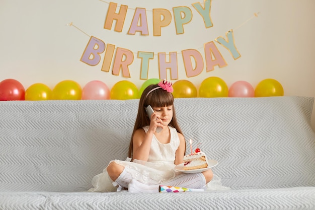 Captura interior de una niñita de cabello oscuro con vestido blanco sentada en un sofá y hablando por teléfono, recibe felicitaciones por su cumpleaños, sosteniendo un pastel con velas encendidas.