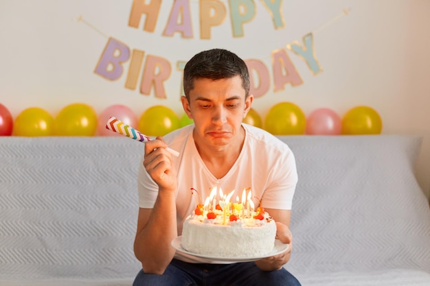 Captura interior de un triste joven caucásico adulto celebra su cumpleaños solo en la sala de estar, soplando las velas en el pastel, sosteniendo el cuerno de fiesta en las manos, expresando tristeza.