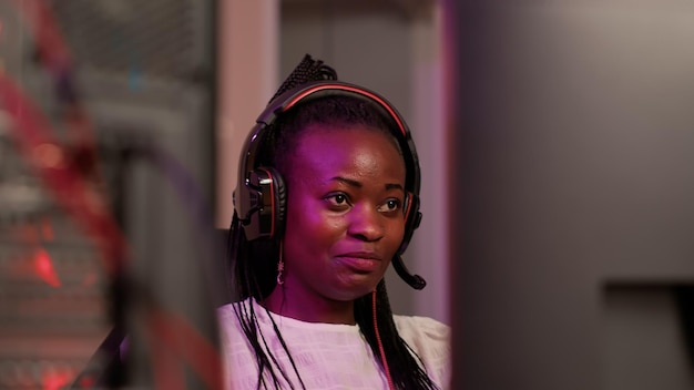 Captura de mano de una jugadora afroamericana con una configuración de PC profesional usando auriculares para juegos hablando con los miembros del equipo. Mujer mirando la pantalla de la computadora mientras juega un juego multijugador en línea.