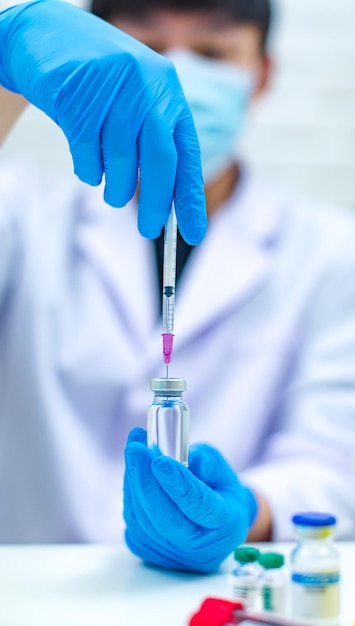 Captura de pantalla de la aguja de la jeringa que succiona la vacuna coronavirus covid19 de un frasco de vial de vidrio para inyección por un científico profesional asiático con bata blanca de laboratorio y máscara facial en un fondo borroso.