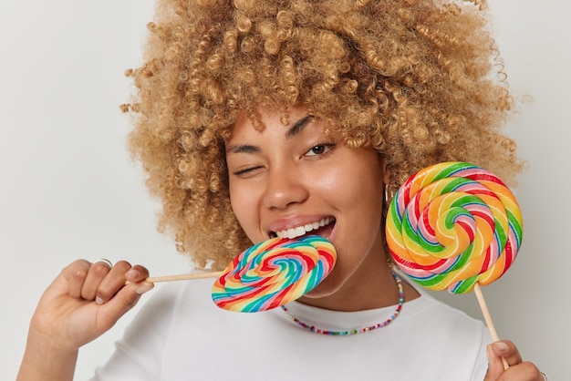Captura de pantalla de una hermosa mujer de cabello rizado que muerde caramelos multicolores en un palo guiña el ojo con una expresión alegre que disfruta comiendo deliciosas piruletas de arco iris dulces posa contra fondo blanco