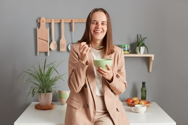 Captura de pantalla de una joven adulta sonriente y riendo con traje beige parada cerca de la mesa en la cocina de casa mirando la cámara y riéndose comiendo un plato sabroso