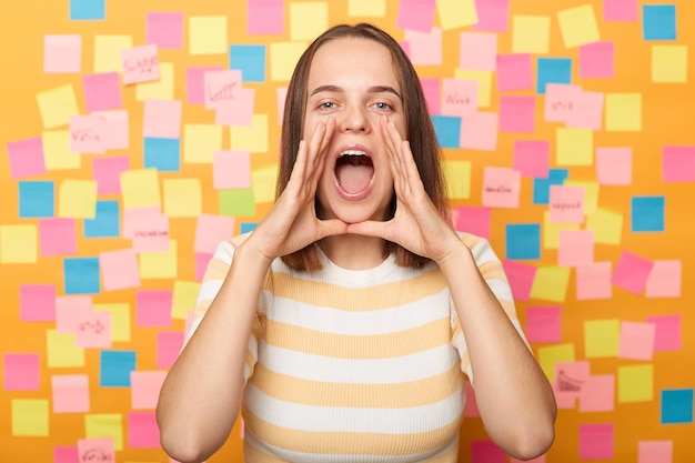 Foto captura de pantalla de una mujer caucásica emocionada y atractiva que usa una camiseta a rayas parada contra una pared amarilla cubierta con pegatinas mantiene las manos cerca de la boca gritando haciendo un anuncio