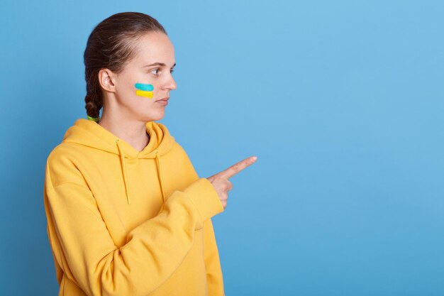 Captura de pantalla de una mujer caucásica seria con capucha y bandera ucraniana en las mejillas posando aislada sobre un fondo azul que señala el camino que muestra el espacio para la publicidad