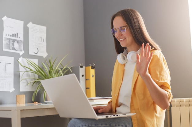 Captura de pantalla de una mujer independiente o estudiante sonriente y encantada con el pelo castaño y una camisa amarilla posando en la oficina con una videollamada en una laptop saludando con la mano