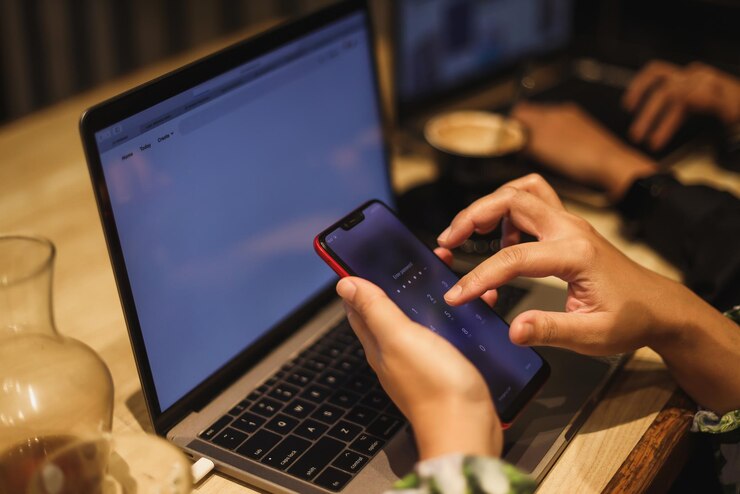 Foto captura de pantalla de una mujer no identificable que usa un teléfono móvil y una computadora portátil en el fondo.