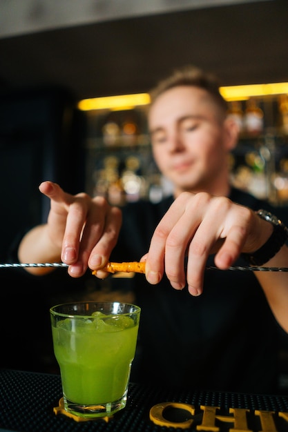 Captura de pantalla vertical de un joven barman exprimiendo un trozo de cáscara de naranja con paja y decorando un cóctel, parado detrás del mostrador del bar en una discoteca moderna con un interior oscuro.