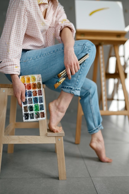 Captura recortada de una artista descalza sosteniendo herramientas de dibujo, paleta de pinturas de acuarela mientras se sienta en una silla en el espacio de trabajo de arte en casa
