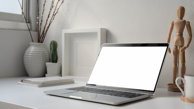 Captura recortada de un lugar de trabajo mínimo con computadora portátil con pantalla en blanco, figura de madera y decoraciones