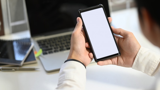 Captura recortada de la mano del empresario recortar smartphone negro con pantalla en blanco en blanco en el moderno escritorio con borrosa portátil y equipo de oficina como