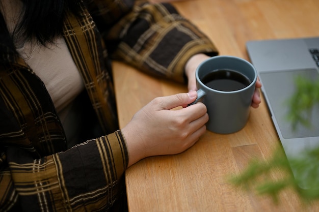 Captura recortada Una mujer con camisa de franela sosteniendo una taza de café sentada en el escritorio de su oficina